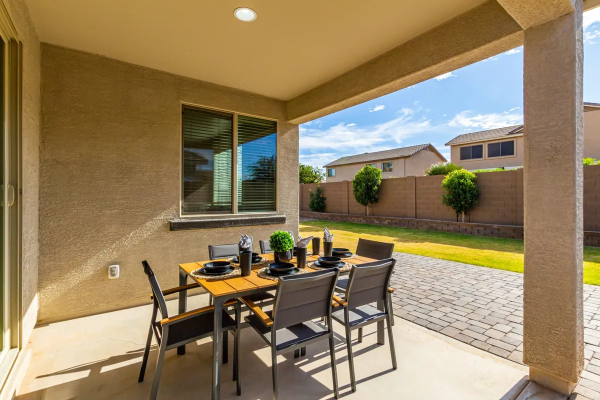 This image presents a welcoming covered patio area with a dining table and chairs set for alfresco meals. Lush green plants line the patio, and the backyard is visible beyond, showing ample space for outdoor activities and relaxation.