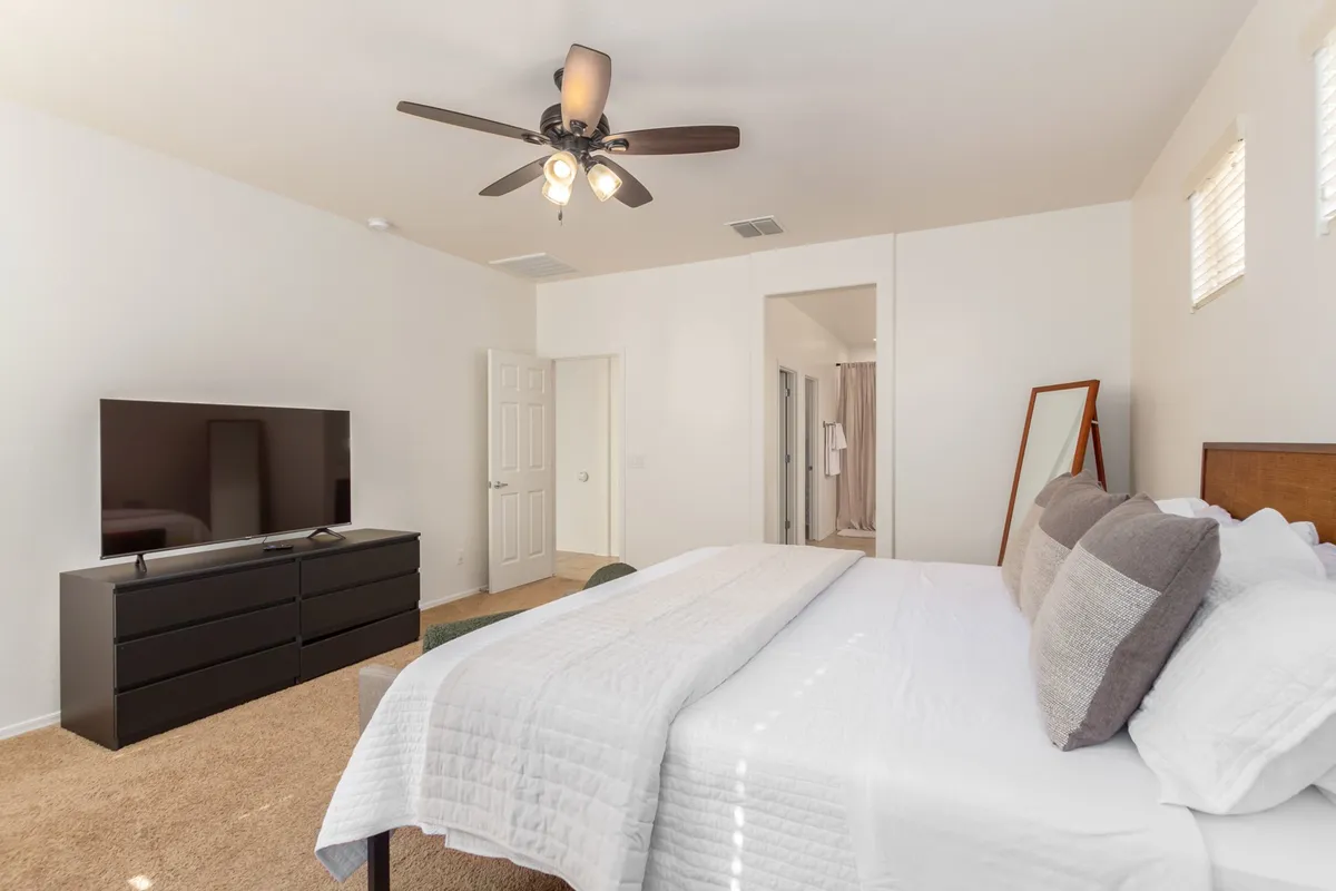 A tranquil bedroom scene featuring a well-made bed, wall-mounted television, dresser, and mirror, all bathed in soft natural light. The overall vibe is restful and conducive to both relaxing and working, with ample surfaces for setting up a laptop or taking video calls.