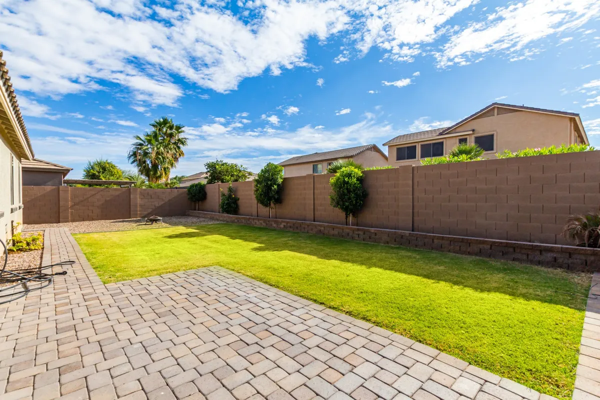 A broad, lush green lawn stretches out, bordered by a paved patio and surrounded by privacy walls and greenery. A palm tree and glimpses of neighboring homes evoke a serene, community-friendly atmosphere ideal for play and relaxation.
