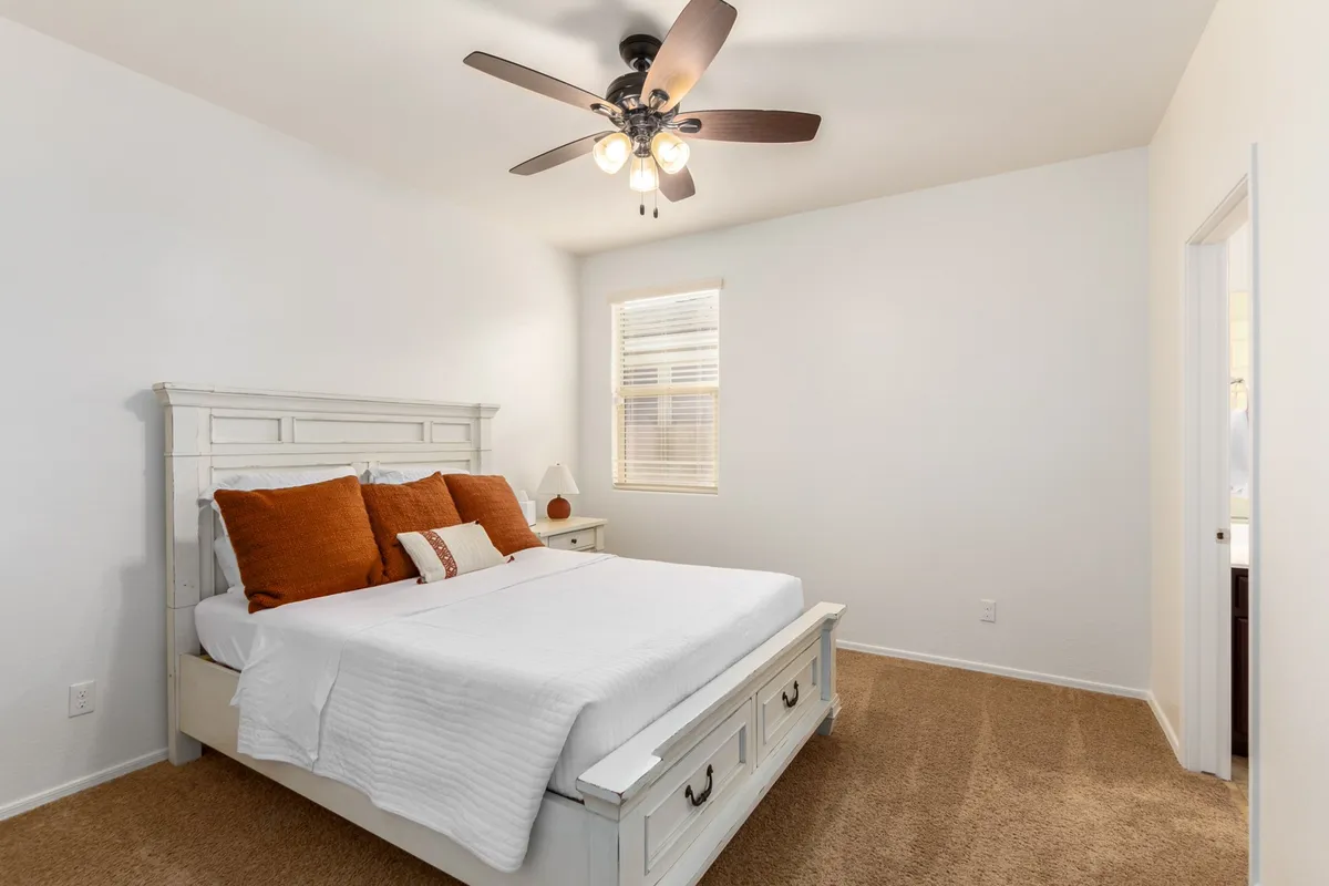 A bright bedroom with a bed dressed in crisp white bedding and vibrant orange pillows sits beneath a ceiling fan, with natural light streaming in through the window. The space feels inviting and peaceful, perfect for unwinding after a busy day.